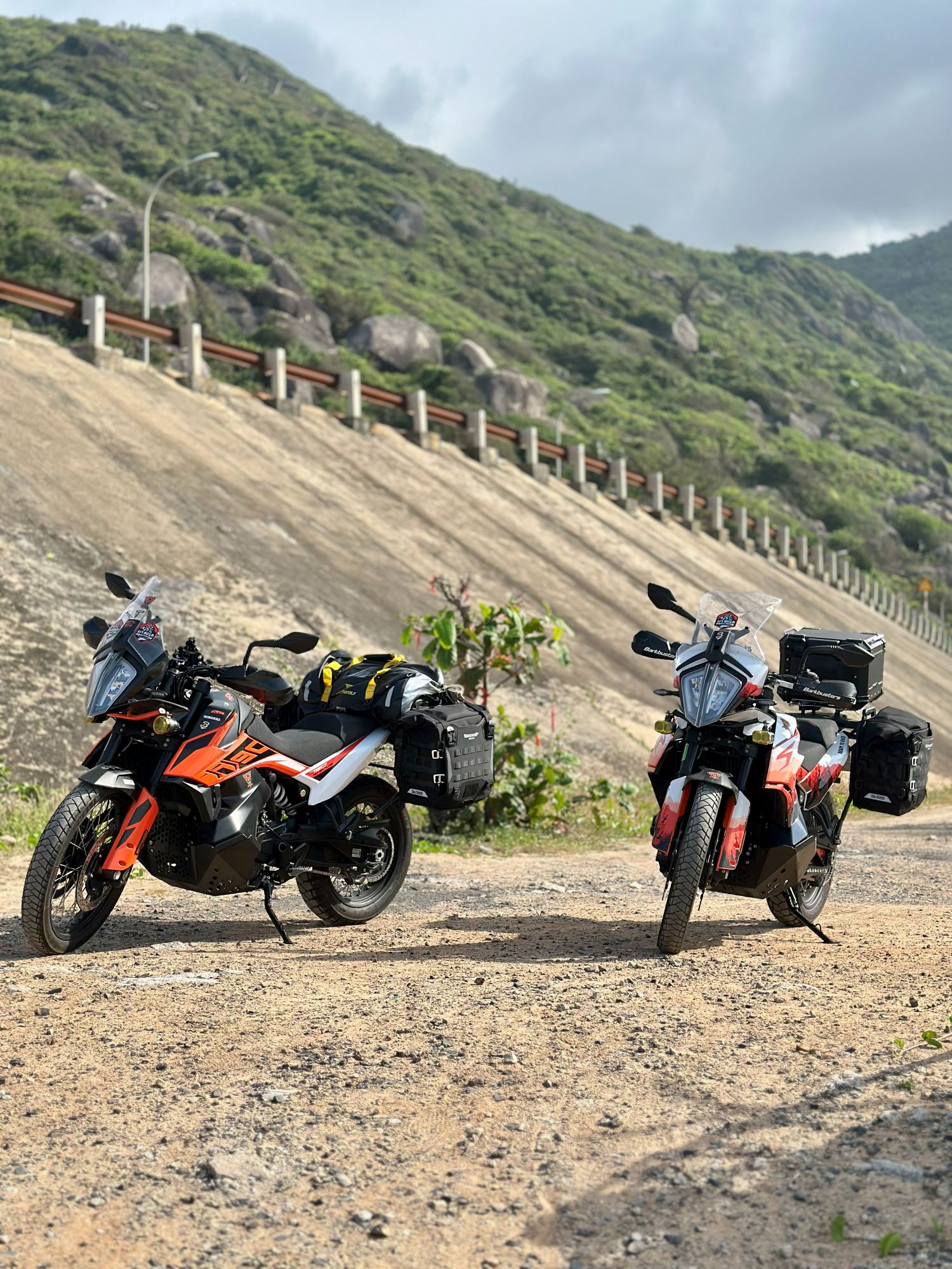 Two loaded KTM 790 Adventures parked below a coastal hill