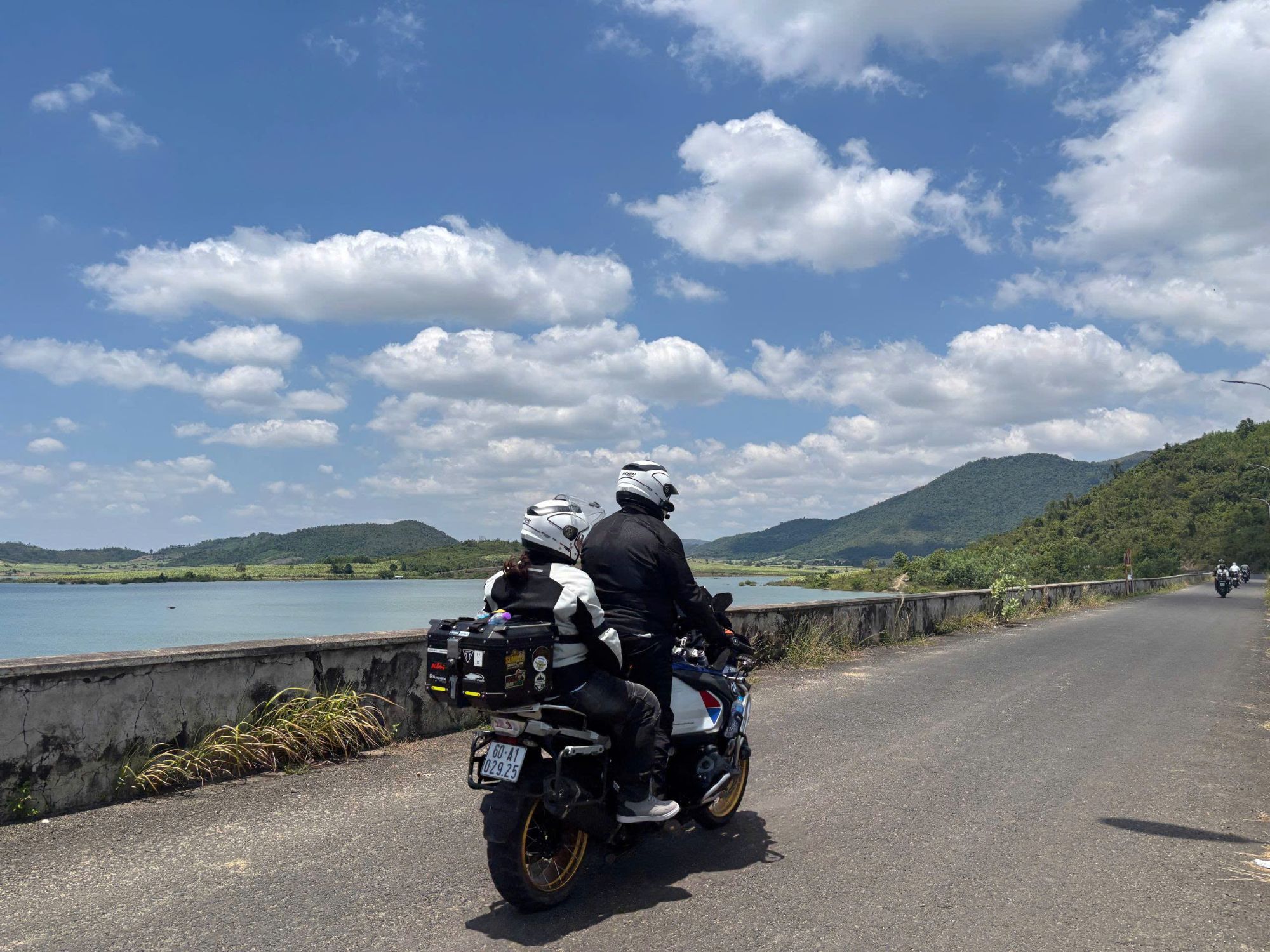 Rider and passenger on a lakeside dam road under a cloudy blue sky