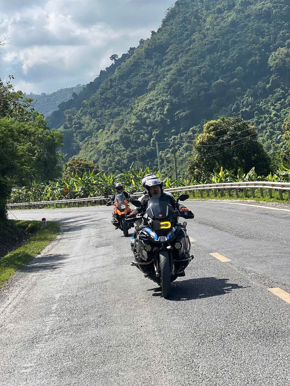 Two riders rounding a bend below a jungle-covered mountain