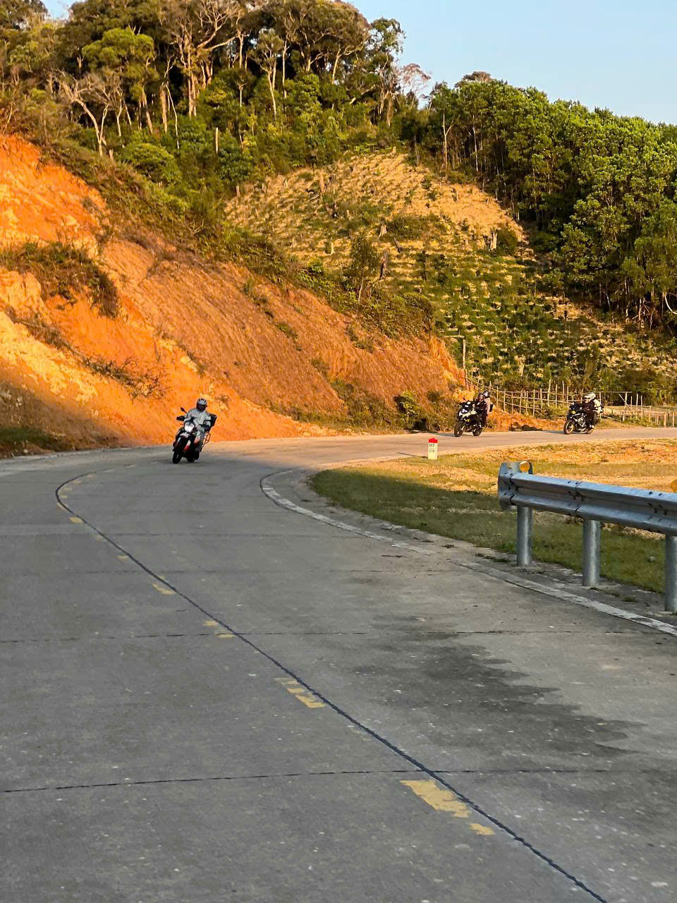 Riders on a curving road past red-earth hills at sunset