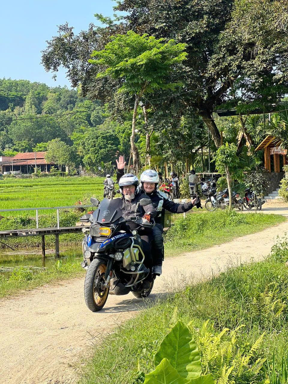 Rider and passenger waving on a dirt lane beside rice fields