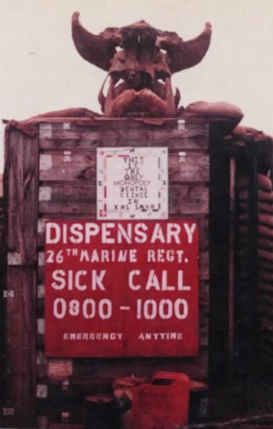 A dispensary sign for the 26th Marine Regiment with sick call hours, topped by a carved devil figure
