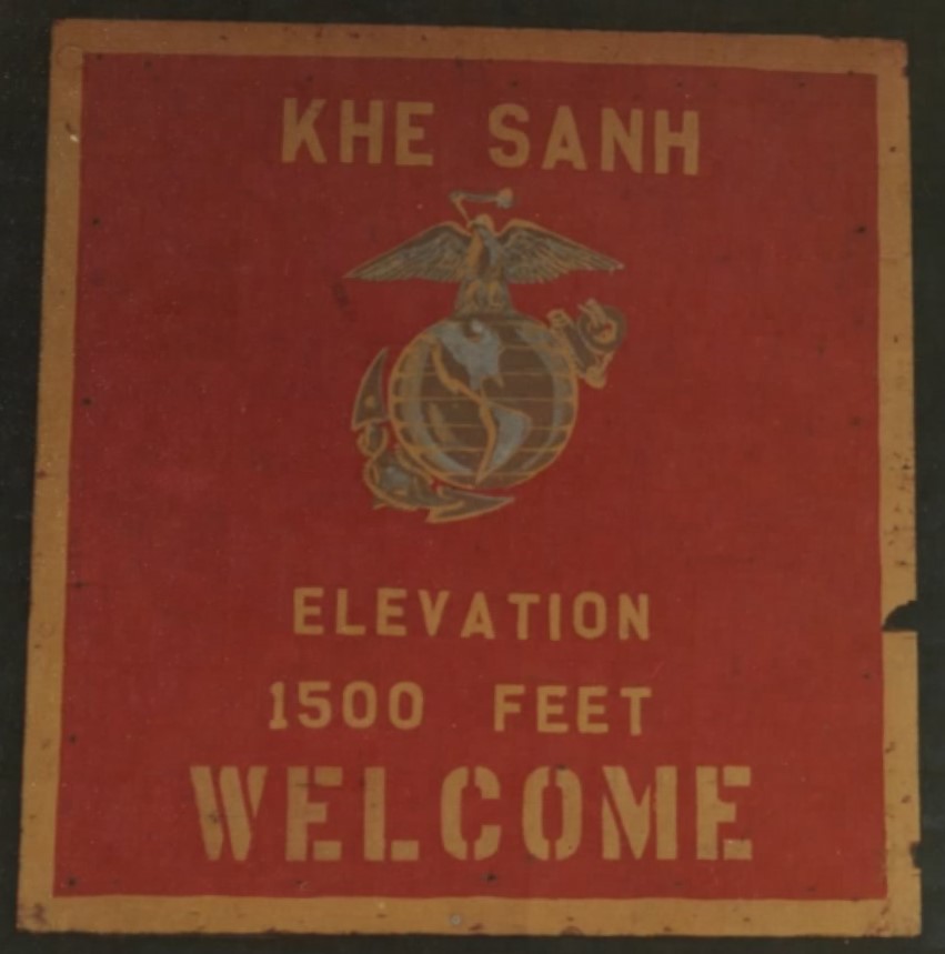 Red sign with the Marine Corps emblem reading Khe Sanh, elevation 1500 feet, welcome
