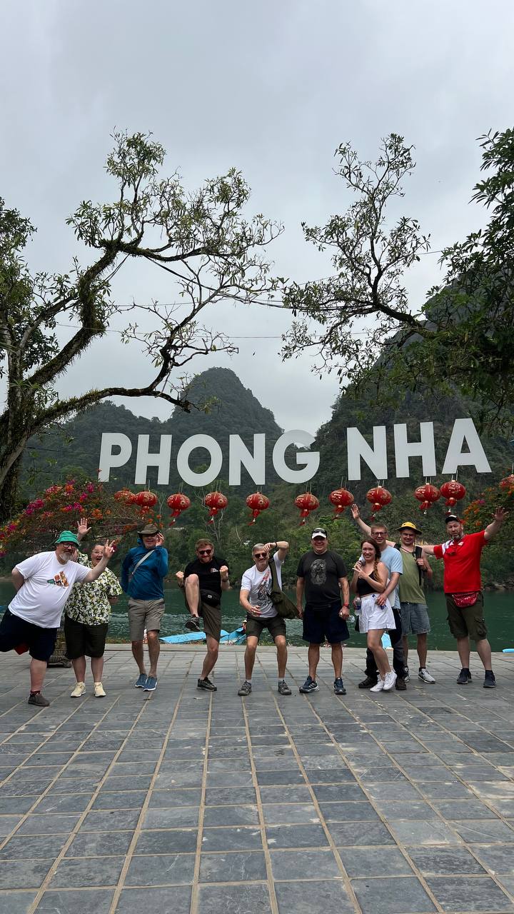 Райдеры DME радуются у большой вывески Phong Nha у реки, позади красные фонари и известняковые вершины